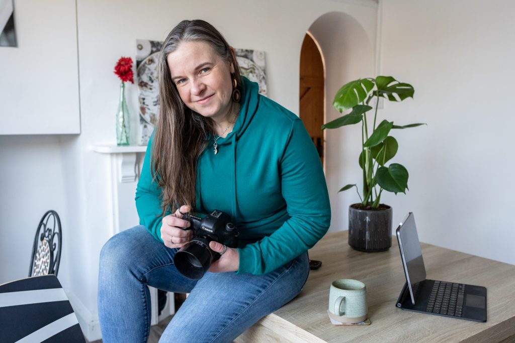 Female photographer in teal hoodie and jeans, with long dark hair, sitting on a table in a lovely, like and attractive room. She's holding her camera. There's a laptop, mug and leafy green plant on the table. The room is a flexible studio space and is set up for a brand photoshoot. North Somerset. Bristol.
